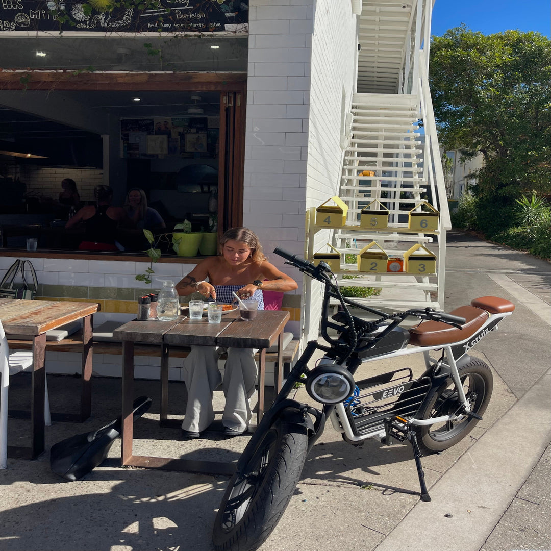Person sitting at an outdoor cafe table with a parked electric bike with pillion seat.